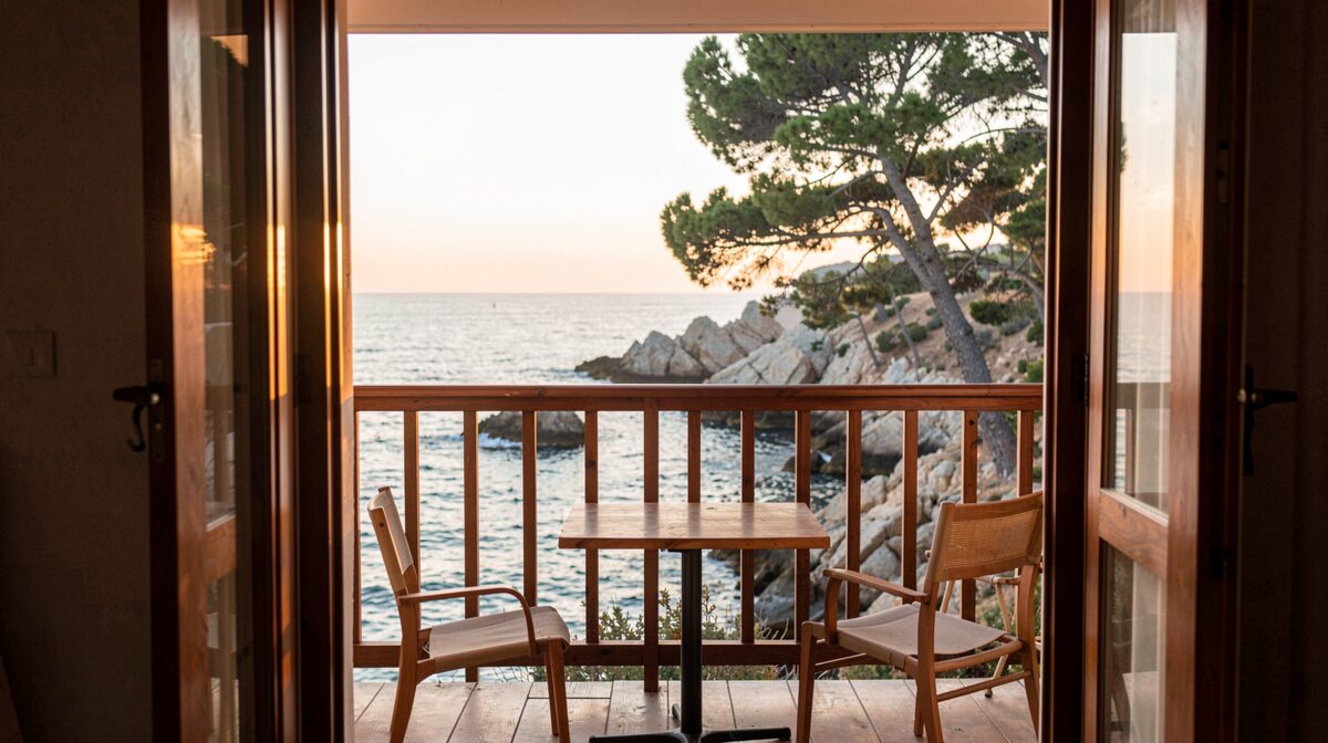 Un balcon d'hôtel en bois face à la Méditerranée au lever du jour, sur la côte varoise, avec une table et deux chaises, la lumière rasante du matin sur l'eau et les pins au-dessus des rochers en arrière-plan, cadrage vertical depuis l'intérieur de la chambre.